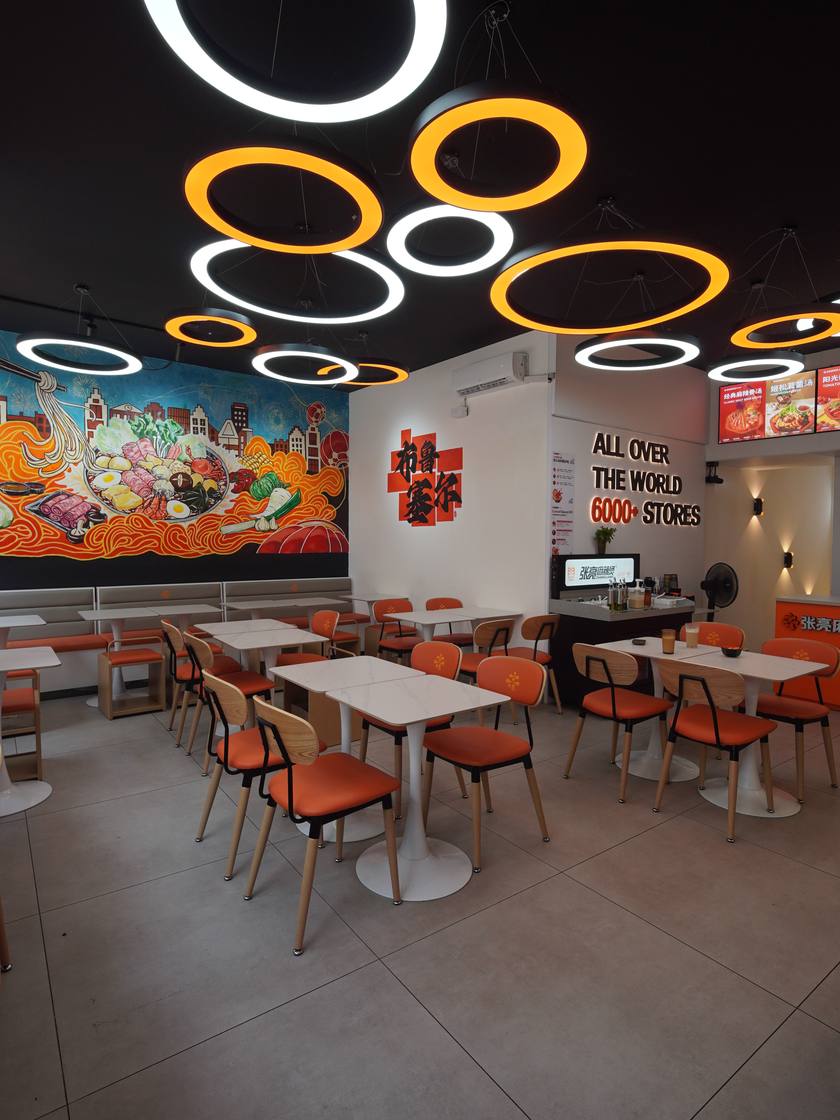 La salle : tables blanches, chaises orange et anneaux lumineux au plafond, devant la fresque murale.